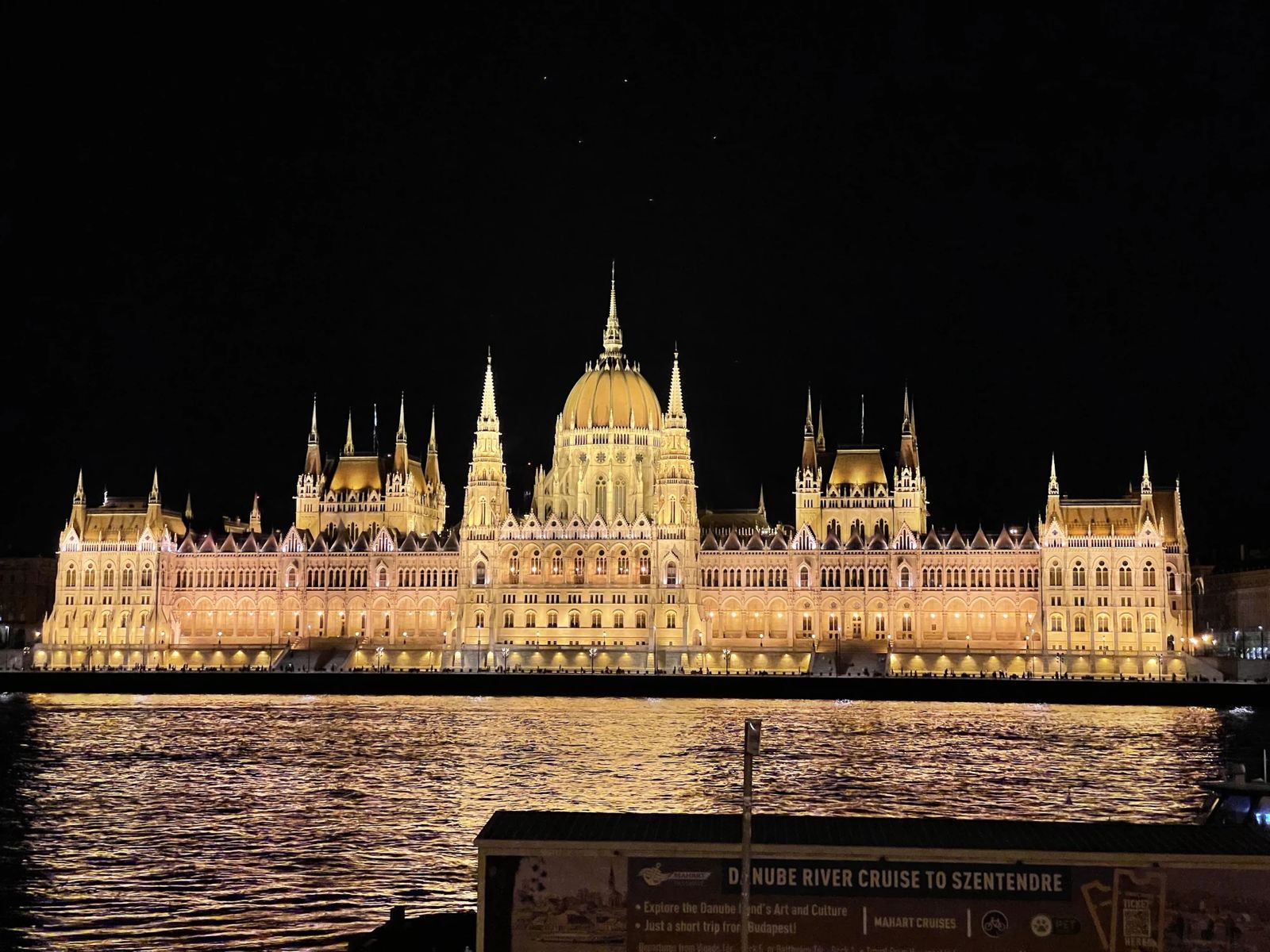 The Hungarian Parliament lit gold at night across the Danube, its lights reflected in the water. A river-cruise sign stands in the foreground.