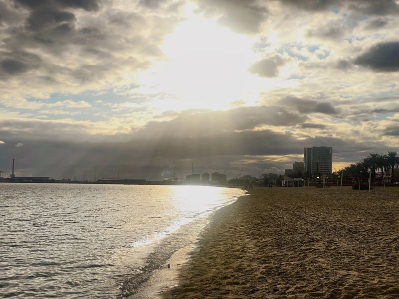 Sun breaks through heavy clouds over a calm bay and a sand beach. A tower block and palm trees stand at the right.