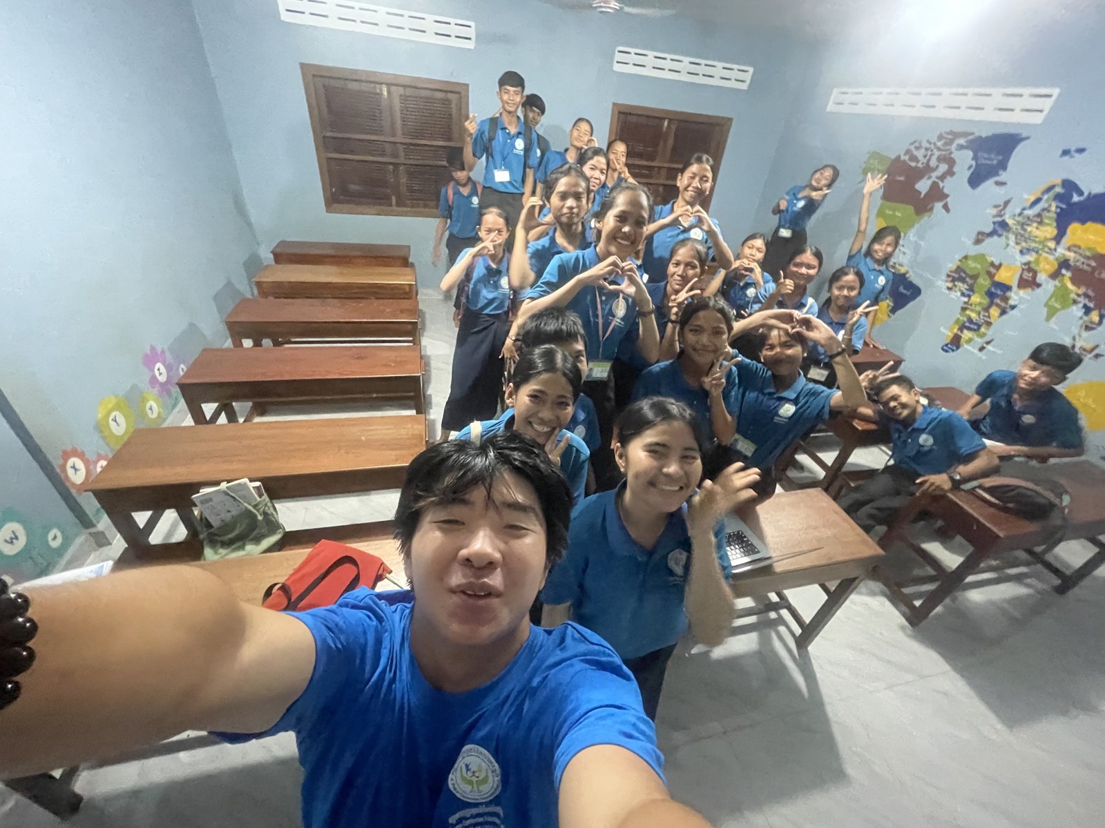 Jonathan takes a selfie at the front of a classroom. About two dozen students in blue PCEF polos stand behind him.