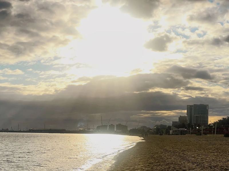 Sunlight breaks through heavy clouds over a bay and a sandy beach, buildings on the far shore.